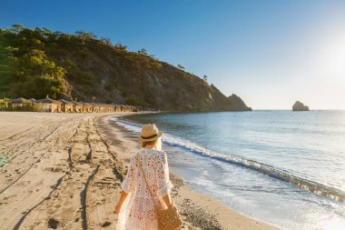 Hasır şapkalı ve beyaz dantelli kimonolu bir kadının kumlu bir plajda yürüdüğünü güneşli bir denize doğru saman şemsiyelerle dizilmiş bir kadın ve tenha bir tropikal tatil kavramını temsil eden bir uçurum.