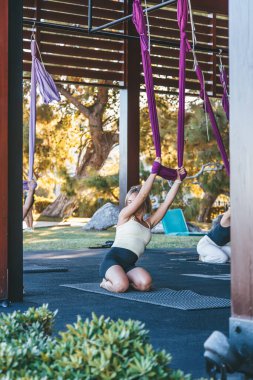 Sarışın bir kadın, bahçedeki bir spor salonunda mor havadan yoga hamakları tutarak sağlığını ve sağlıklı yaşam tarzını temsil eden bir yerde diz çöküyor.
