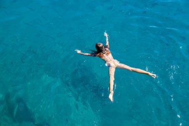 Kolları ve bacakları kristal berraklığında yüzen genç bir kızın yüksek açılı çekimi. Turkuaz mavi su, yaz tatili sırasında özgürlük ve sakinlik hissi veriyor.