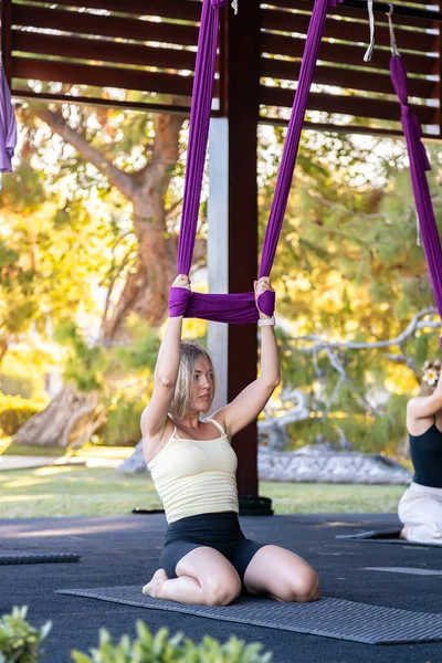 Sinek yogası sırasında mindere diz çökmüş zinde bir genç kadın. İki eliyle canlı bir mor ipek hamağa tutunuyor, esneme ve nefes alışına odaklanıyor.