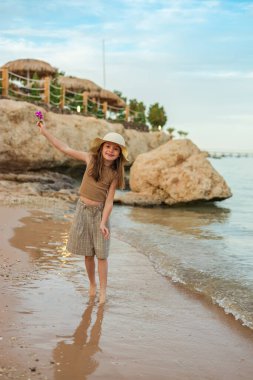 Saman şapkalı, elinde mor bir çiçekle, tropikal bir seyahat ve yaz tatili sırasında kumlu deniz kıyısında çıplak ayakla yürürken mutlu küçük kız.
