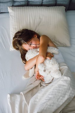 Rahat bir yatakta uzanmış genç bir kızın yukarıdan aşağıya bakışı, büyük, yumuşak bir tavşan oyuncağına sarılırken nazikçe gülümsemesi. Görüntü, huzurlu bir sabah rahatlığı ya da dinlendirici bir öğleden sonra uykusunu yakalar. Çocukluk sağlığı için ideal bir ortam, aile yaşam tarzı ya da...