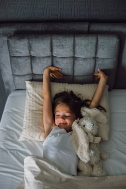 Konforlu bir tavşan oyuncağıyla rahat bir yatakta uzanan mutlu bir genç kız, evde huzurlu bir sabahın tadını çıkarıyor ya da çocukluk sağlığı ya da günlük bir konsept için tatilde.