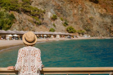 Beyaz dantelli tunik ve hasır şapka giyen, tahta korkuluklara yaslanan ve güzel turkuaz bir körfeze bakan bir kadının arka görüntüsü. Lüks bir Akdeniz tatili sırasında sessiz bir düşünce anı.