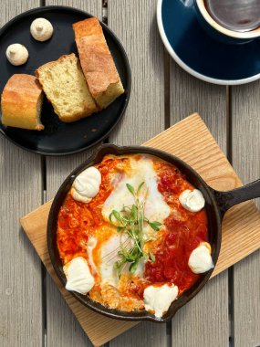 Haşlanmış yumurta, domates sosu, krem peynir ve mikroyeşillik ile birlikte kahvaltıda kabuklu ekmek ve kahveyle servis edilen dökme demir tavada iştah açıcı shakuka.