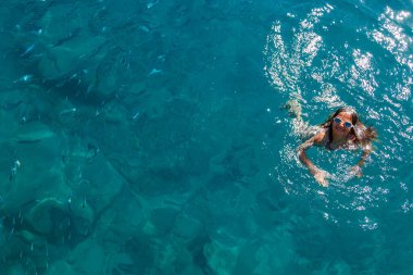 Şeffaf mavi okyanusta yüzen gözlüklü, gülümseyen bir çocuk. Yakınlardaki tropikal bir sahil beldesinde seyahat ve rahatlama konseptinde balık sürüleri.