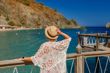 Sahil kenarında dinlenen bir kadın turistin arka görüntüsü. Açık mavi suyu ve dağları olan tropikal bir sahil lagününün manzarası.