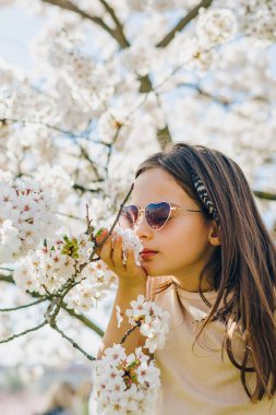 Güneş gözlüklü güzel bir genç kız güneşli bahar parkında beyaz kiraz çiçekleri koklarken doğa bağlantısı ve mevsimsel yaşam tarzı konsepti için