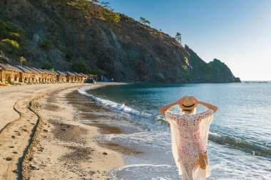 Yaz elbisesi giyen ve örgü ören kadın gezgin lüks villaların yakınındaki tropikal sahillerde gezinti yapıyor ve Akdeniz tatilini ve barışçıl seyahat titreşimlerini temsil eden turkuaz suyu temsil ediyor.