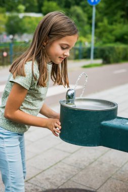 Çağdaş bir mahalledeki vatandaşlar için sağlıklı hidrasyon alışkanlıklarını ve kamu altyapısını gösteren yeşil metal bir su dağıtıcısına susuzluğunu gideren bir çocuk.