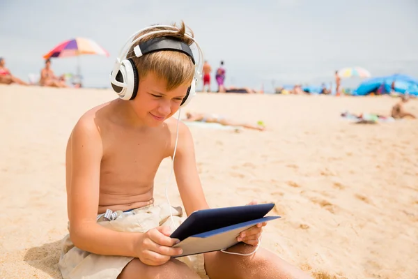 Chłopiec korzysta z komputera typu Tablet, siedząc na plaży — Zdjęcie stockowe