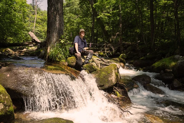 Tourist with a camera sitting on a stone near river — Stock Photo, Image