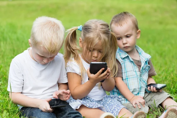 Niños jugando en teléfonos inteligentes sentados en la hierba —  Fotos de Stock