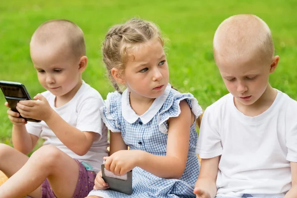 Children using smartphones sitting on the grass — Stock Photo, Image