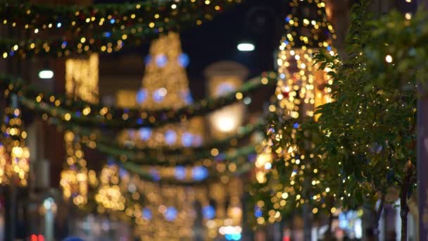 Imagen borrosa de la ciudad europea de Navidad iluminada con luces navideñas. Guirnaldas en árboles y casas parpadeando luces azules y amarillas — Vídeos de Stock