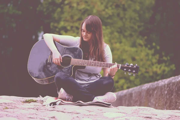 Pretty teenager girl playing guitar while sitting on the stone bridge — Stock Photo, Image