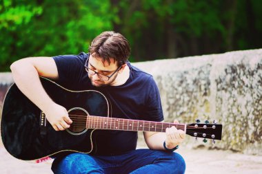 Gitar çalan genç adam köprüde oturuyor.