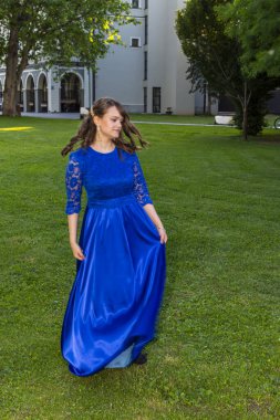 Mezuniyet balosu kızı, modern ve zarif bir mavi elbiseyle dönüyor. Sevimli ve şık kıyafetler. Mezuniyet töreni için doğadan güzel bir fotoğraf.