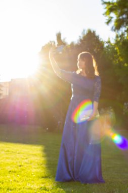 Zarif elbiseli güzel, gülümseyen kadın manken yaz parkında selfie çekiyor. Doğada mavi kıyafetli, sevimli, modaya uygun bir kız. Sanat fotoğrafı. Seçici odak