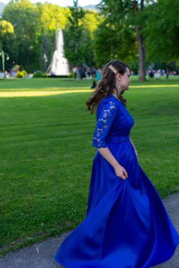 Mezuniyet balosu kızı, modern ve zarif bir mavi elbiseyle dönüyor. Sevimli ve şık kıyafetler. Mezuniyet töreni için doğadan güzel bir fotoğraf.