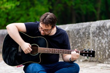 Gözlüklü genç adam eski Roma köprüsünde oturmuş gitar çalıyor.