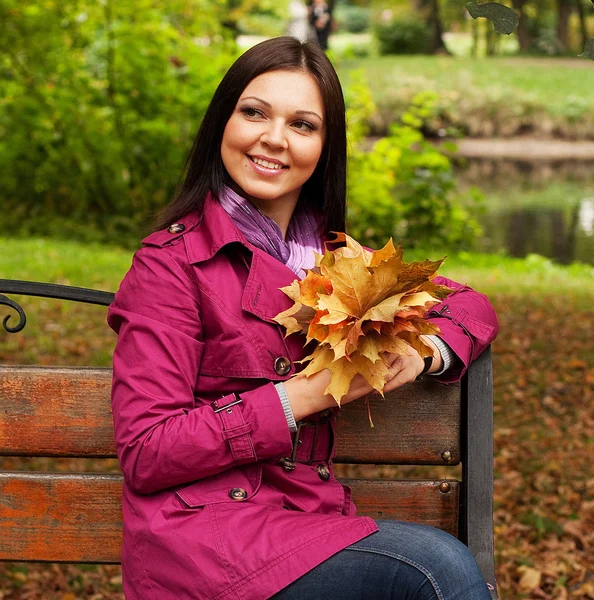 Młoda kobieta z liści jesienią, siedząc na ławce — Zdjęcie stockowe