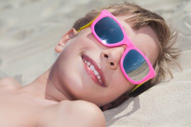 güzel çocuk güneşlenme ve gülümseyerek kumlu bir plaj üzerinde yalan renkli güneş gözlüğü takıyor