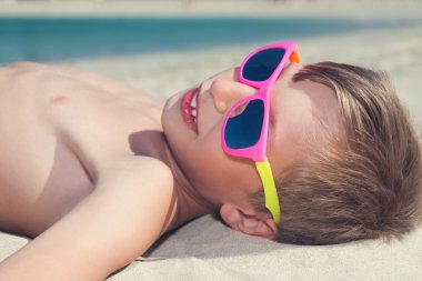komik çocuk bronzlaşma ve gülümseyerek kumlu bir plaj üzerinde yalan renkli güneş gözlüğü takıyor
