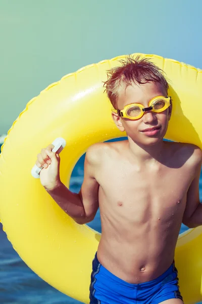 Funny chilld with swimmer goggles standing on beach with yellow float smiling — Stock Photo, Image