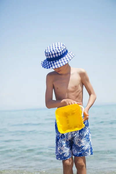 Child in swimmer shorts standing on beach holding a toy bucket pouring water — Stock Photo, Image