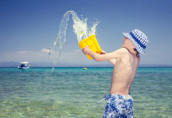 Funny little boy with a panama playing in the sea throwing a yellow toy bucket of water laughing — Stock Photo, Image