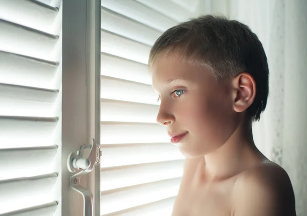 Kleine jongen poseren voor een venster met blinds met een grappige expressie — Stockfoto
