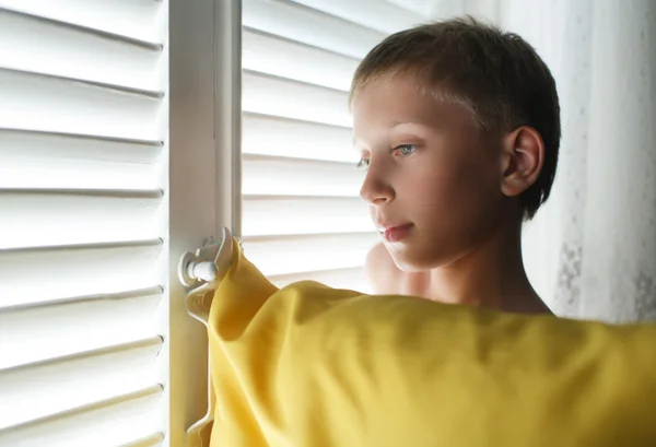 Openhartig portret van een mooi klein kind met een kussen staan door een venster met blinds op zoek triest. — Stockfoto
