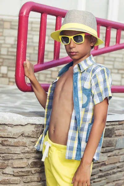 Bellissimo bambino divertente con eleganti occhiali da sole gialli, pantaloncini e cappello in posa all'aperto in estate — Foto Stock