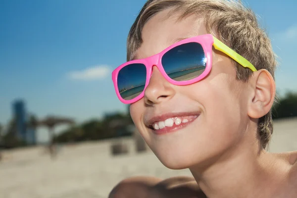 Beatiful child wearing colorful sunglasses on sandy beach tanning and smiling — Stock Photo, Image