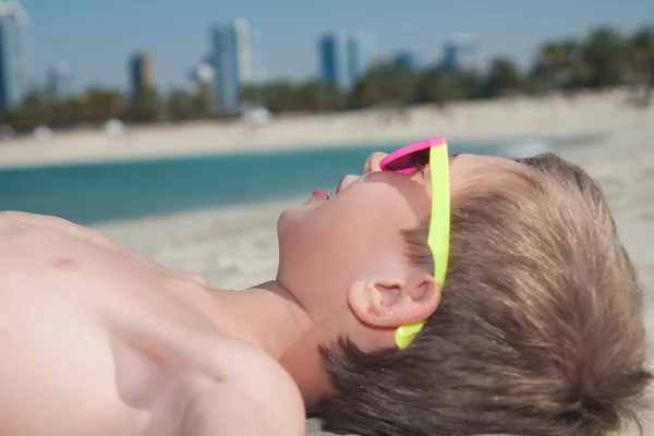 Funny child wearing colorful sunglasses lying on sandy beach sunbathing and smiling — Stock Photo, Image