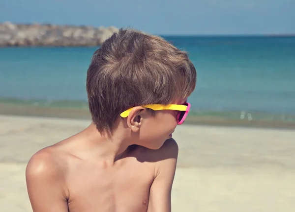 Kind draagt stijlvolle zonnebril looien op zandstrand glimlachen op zoek op zee — Stockfoto