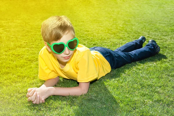 Счастливый ребенок с забавными солнцезащитными очками лежит на зеленой траве в солнечном парке — стоковое фото