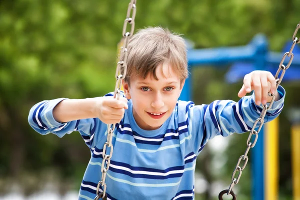 Hermoso niño feliz balanceándose en un parque infantil en un parque soleado y sonriendo . — Foto de Stock