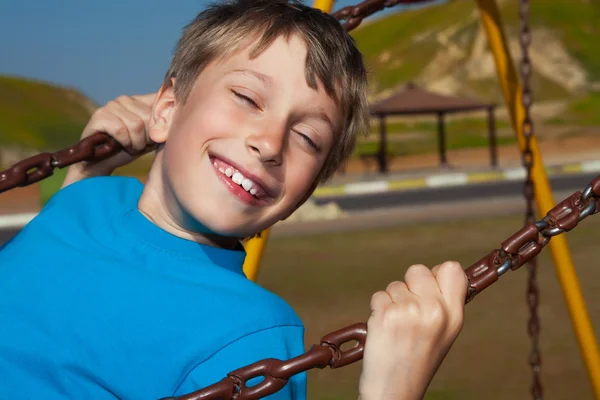 Bonito menino feliz balançando em um playground em um parque ensolarado e rindo — Fotografia de Stock