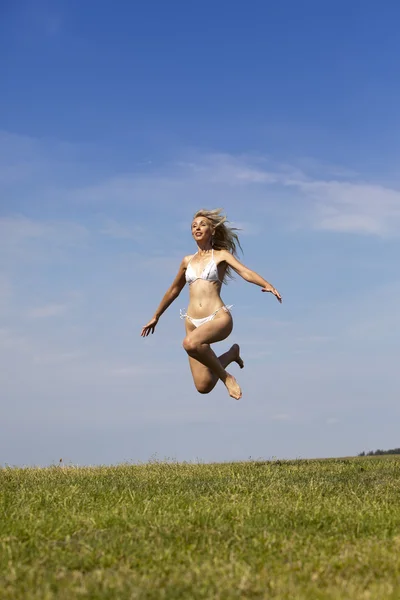 De gelukkige vrouw in witte bikini springt in een zomer groen veld tegen de blauwe hemel — Stockfoto