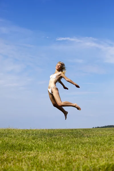 Die glückliche Frau im weißen Bikini springt in einem sommergrünen Feld gegen den blauen Himmel — Stockfoto