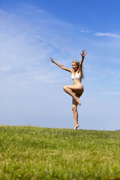 De gelukkige vrouw in witte bikini springt in een zomer groen veld tegen de blauwe hemel — Stockfoto