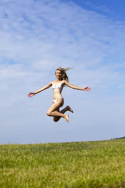 La mujer feliz en bikini blanco salta en un campo verde de verano contra el cielo azul —  Fotos de Stock