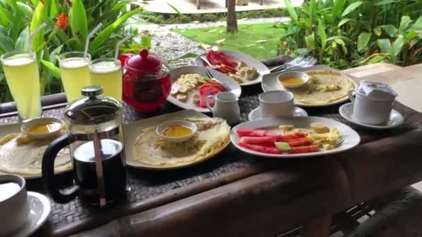 Pequeno-almoço, panquecas e frutas, é servido no terraço exterior, com vista para as plantas tropicais e. Indonésia — Vídeo de Stock