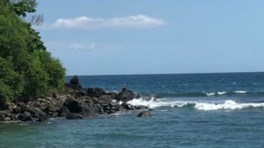 Kumsalda dalgalar çarpar, tropikal kumsalda deniz sörfü yapar balıkçılar oltayla balık yakalar.