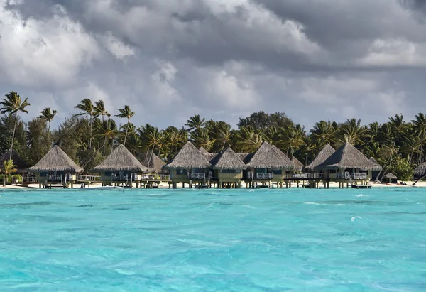 Tipik Polinezya manzarası - Palmiye ağaçları ve su üzerinde küçük evleri olan deniz manzarası — Stok fotoğraf