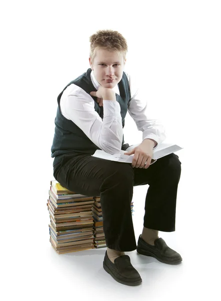 The schoolboy in a school uniform sits on a pack of books, with the opened book in hands — Stock Photo, Image