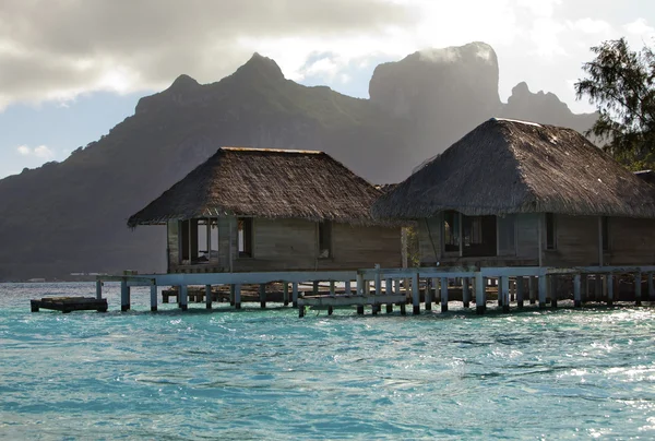 Le capanne buttate distrutte su acqua e isola con palme nell'oceano e montagne su uno sfondo — Foto Stock