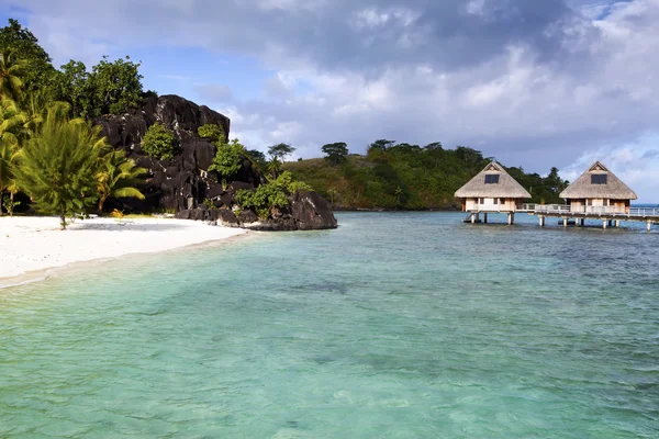 Paisaje polinesio típico: isla con palmeras y casitas sobre el agua en el océano y montañas sobre un fondo —  Fotos de Stock